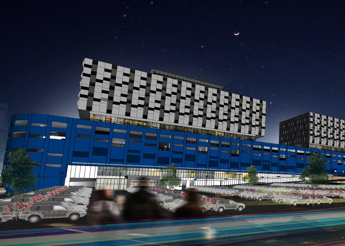 A digital rendering of a modern industrial complex at night, featuring a prominent blue building with a white patterned tower and other connected structures, set against a dark sky with light trails from cars in the foreground.