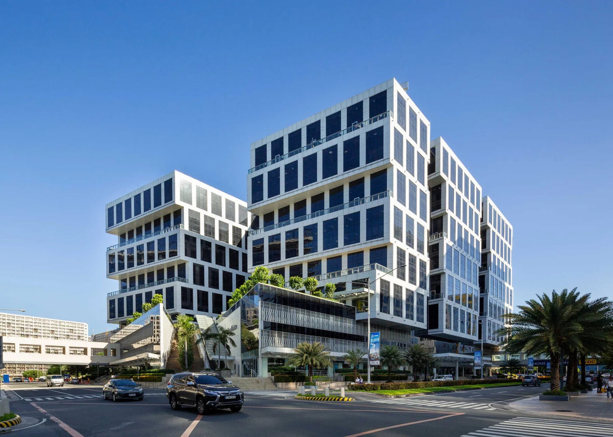 Modern multi-story office buildings with white facades and dark glass windows, situated on a city street under a clear blue sky with palm trees and a few cars in the foreground.