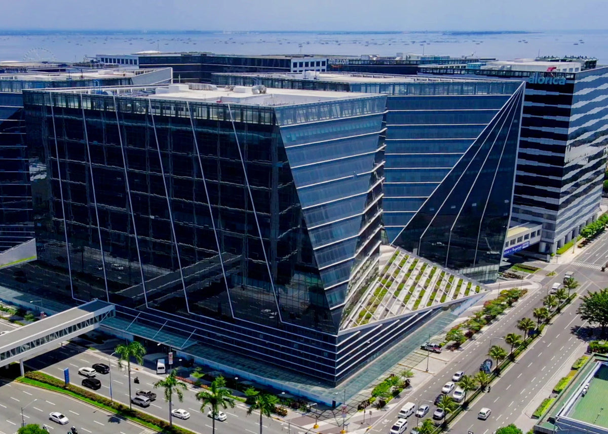 Aerial view of a modern glass office building complex with angular geometric forms, surrounded by roads, palm trees, and water in the distance.