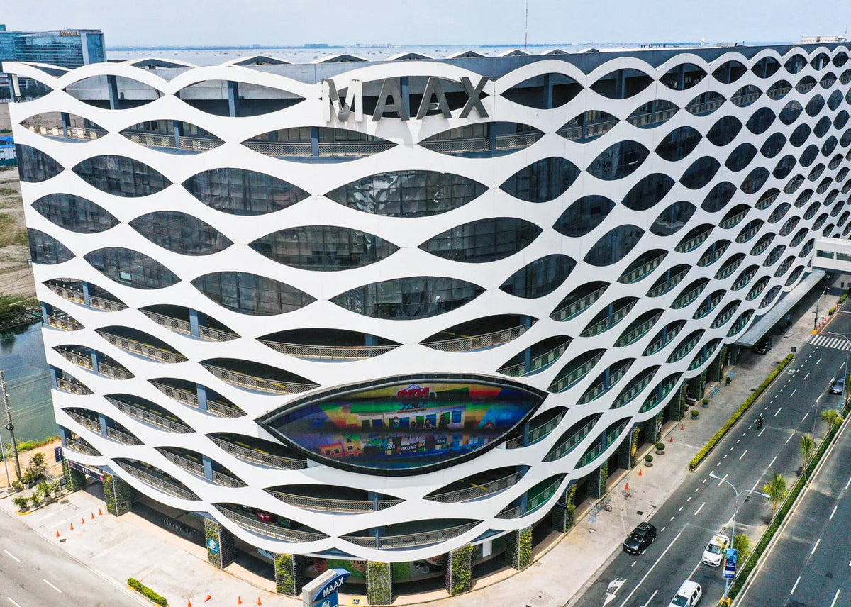 Aerial view of a modern white building with a distinctive honeycomb-like wave pattern facade, featuring a large colorful oval mural near the ground level and a MAXX sign on top, surrounded by roads and greenery.