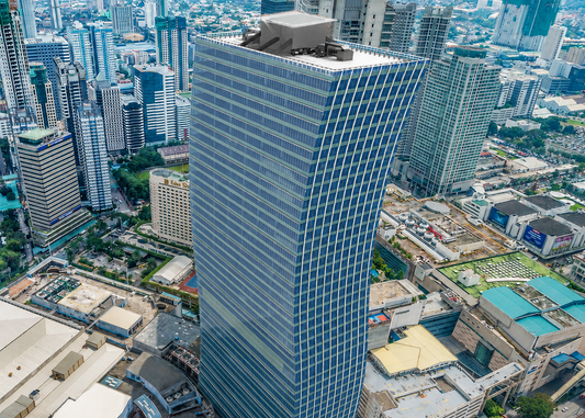 An aerial view of a tall modern glass skyscraper with a curved, tapered design, situated in a dense urban city environment with surrounding buildings, roads, and greenery.