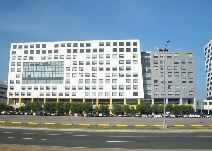 A large modern white office building with many windows, set behind a road with parked cars and a row of trees, under a clear blue sky.