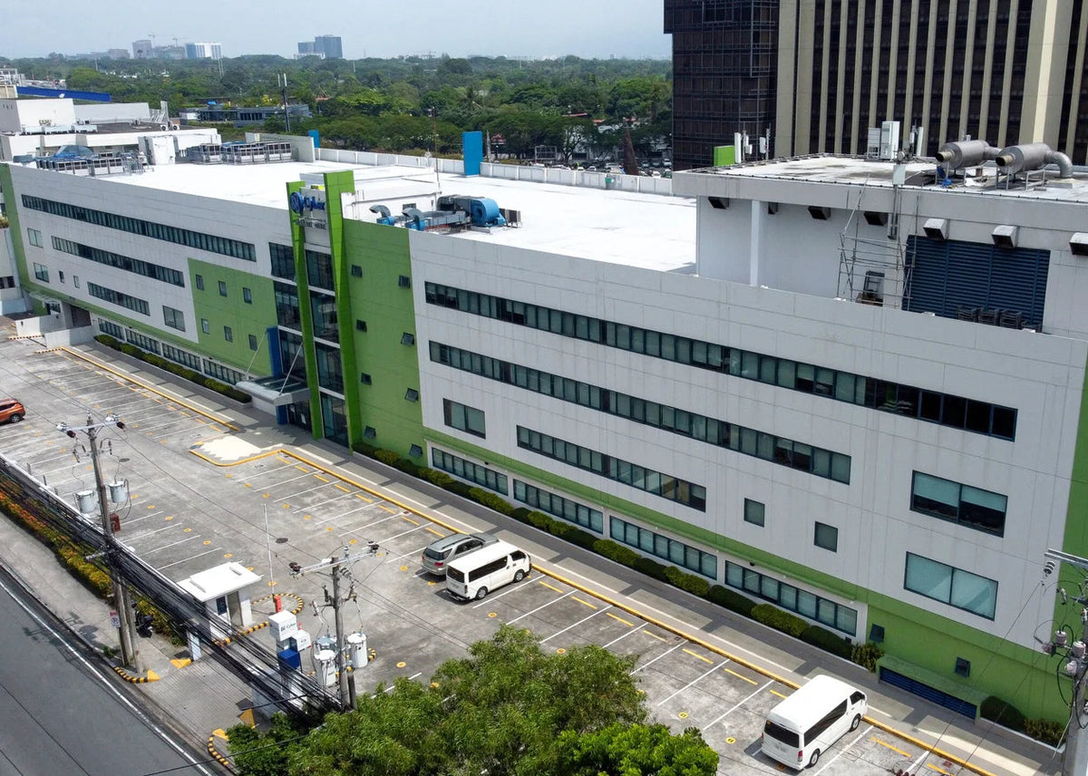 Aerial view of a large modern white and green industrial building with a parking lot, surrounded by other buildings and trees.