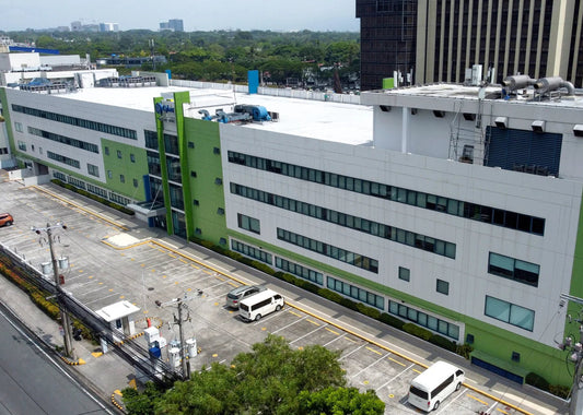 Aerial view of a large modern white and green industrial building with a parking lot, surrounded by other buildings and trees.