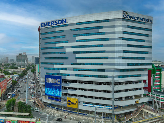 Aerial view of a large modern office building with prominent 'EMERSON' and 'CONCENTRIX' signage on the exterior, surrounded by streets, vehicles, and nearby structures under a blue sky.