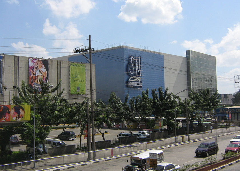 SM City Fort Bonita shopping mall exterior under a blue sky with clouds, featuring a large blue and gray facade, trees in the foreground, and a busy road with cars below.