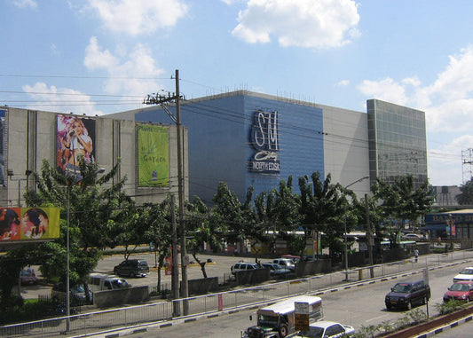 SM City Fort Bonita shopping mall exterior under a blue sky with clouds, featuring a large blue and gray facade, trees in the foreground, and a busy road with cars below.