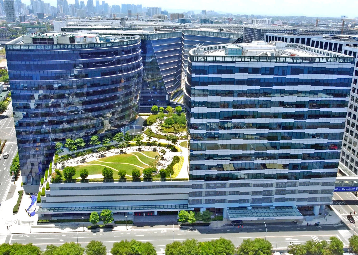 Aerial view of a modern office building complex with two curved towers, a central landscaped green courtyard, and surrounding city streets and skyline.