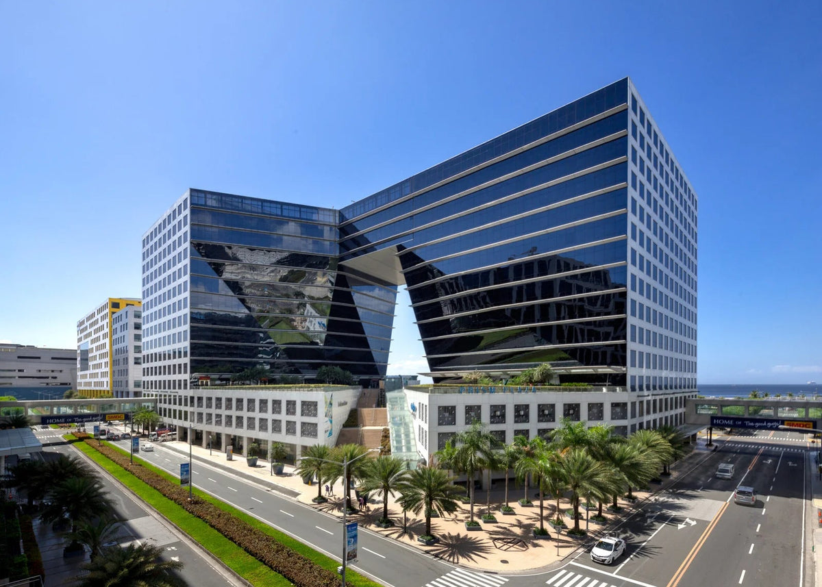 A modern glass office building with a distinctive angular design, featuring a central glass pyramid structure, surrounded by palm trees, a landscaped median, and a busy city street under a clear blue sky.